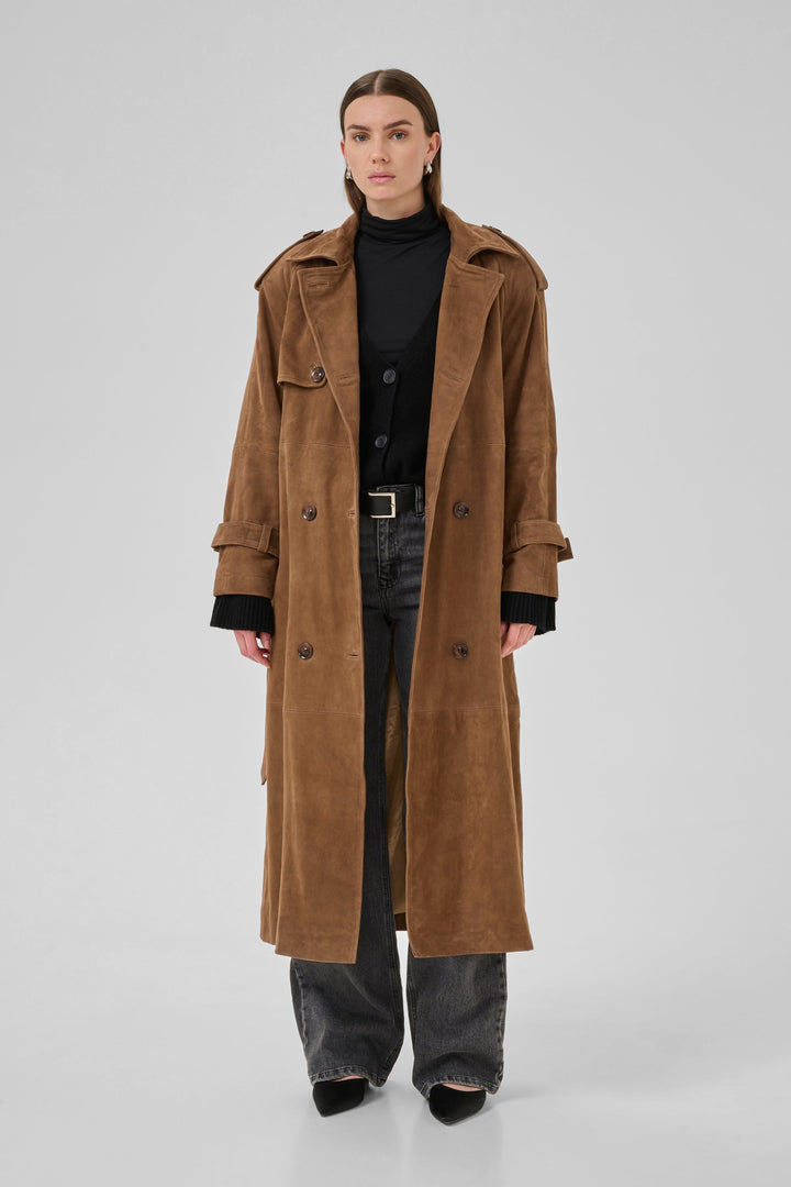 Person iført cognacfarvet trenchcoat med dobbeltknapper, epauletter og bælte over sort tøj.