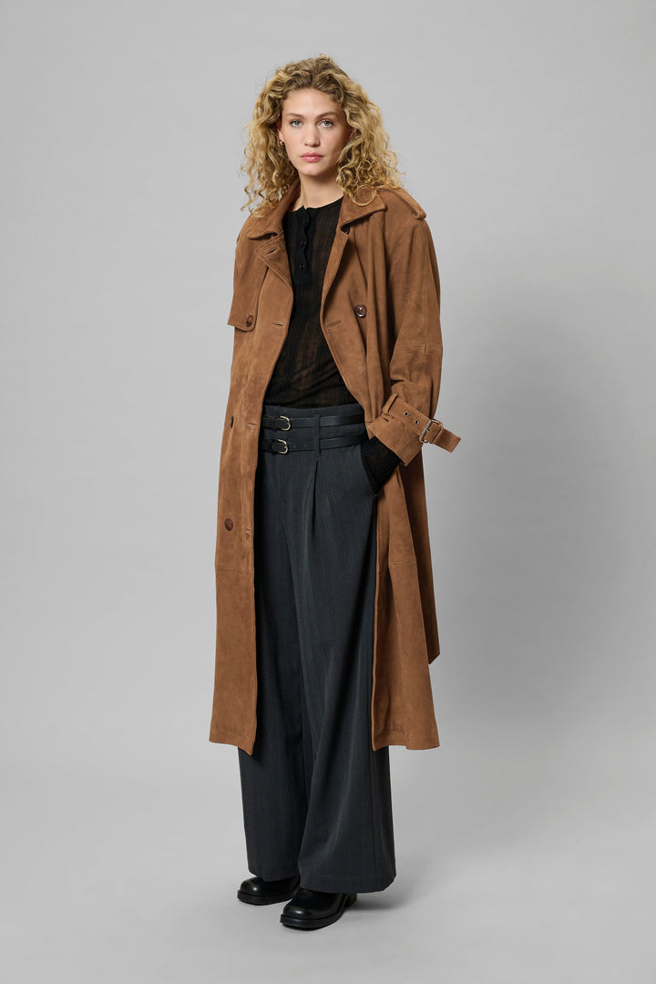 Person iført cognac trenchcoat med dobbeltknapper, epauletter og bælte, sorte bukser og sko.