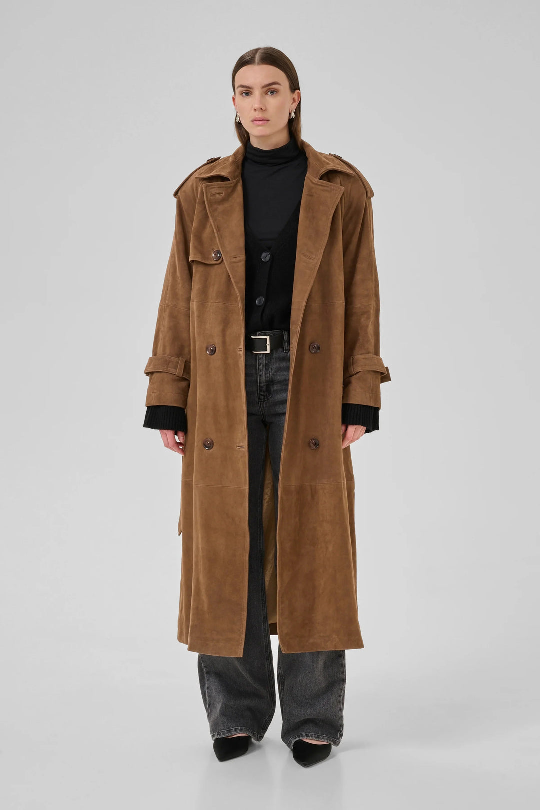Person iført cognacfarvet trenchcoat med dobbeltknapper, epauletter og bælte over sort tøj.