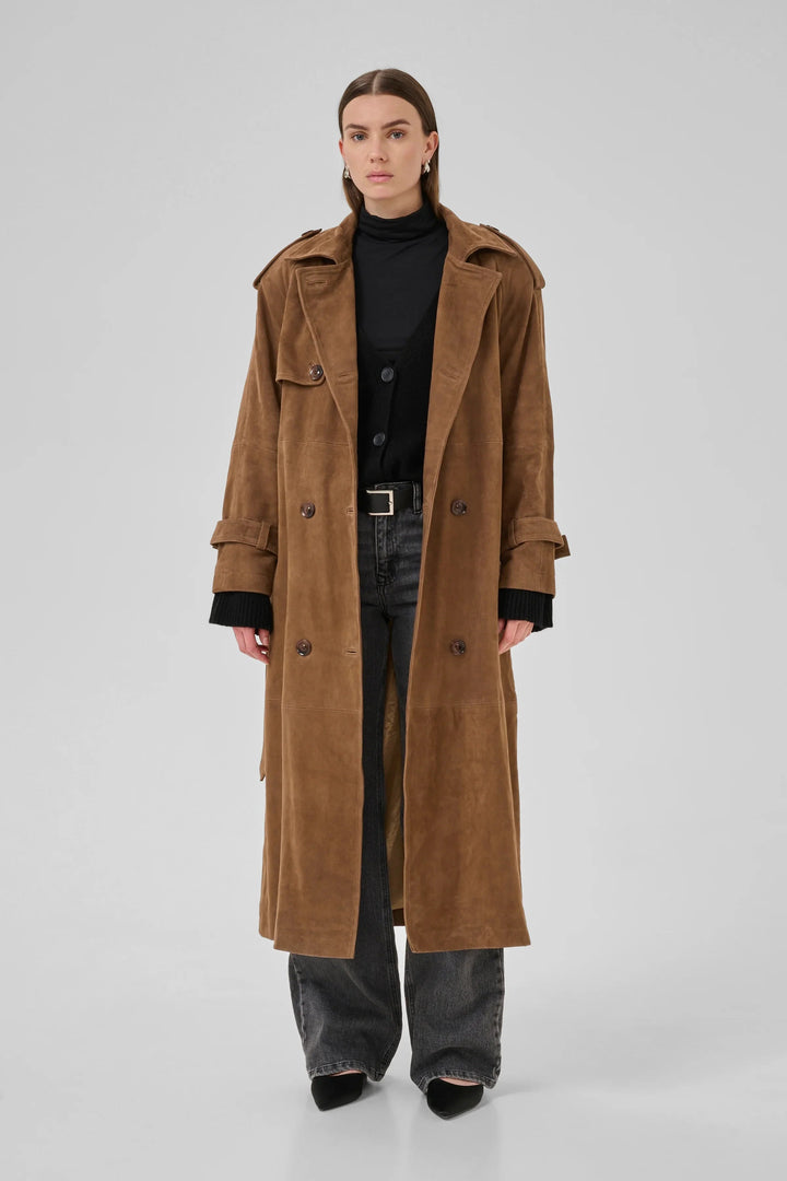Person iført cognacfarvet trenchcoat med dobbeltknapper, epauletter og bælte over sort tøj.