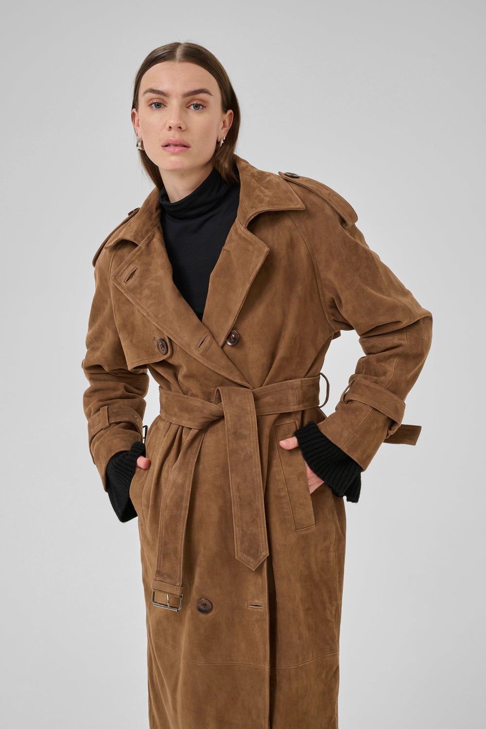 Person i cognac trenchcoat med dobbeltknap, bælte og epauletter, iført sort trøje.