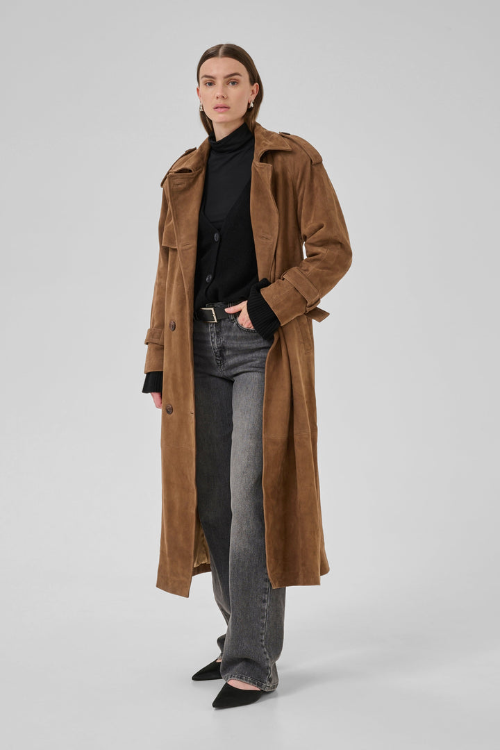 Person iført cognacfarvet trenchcoat med dobbeltknappet front og bælte, sort top og grå jeans.