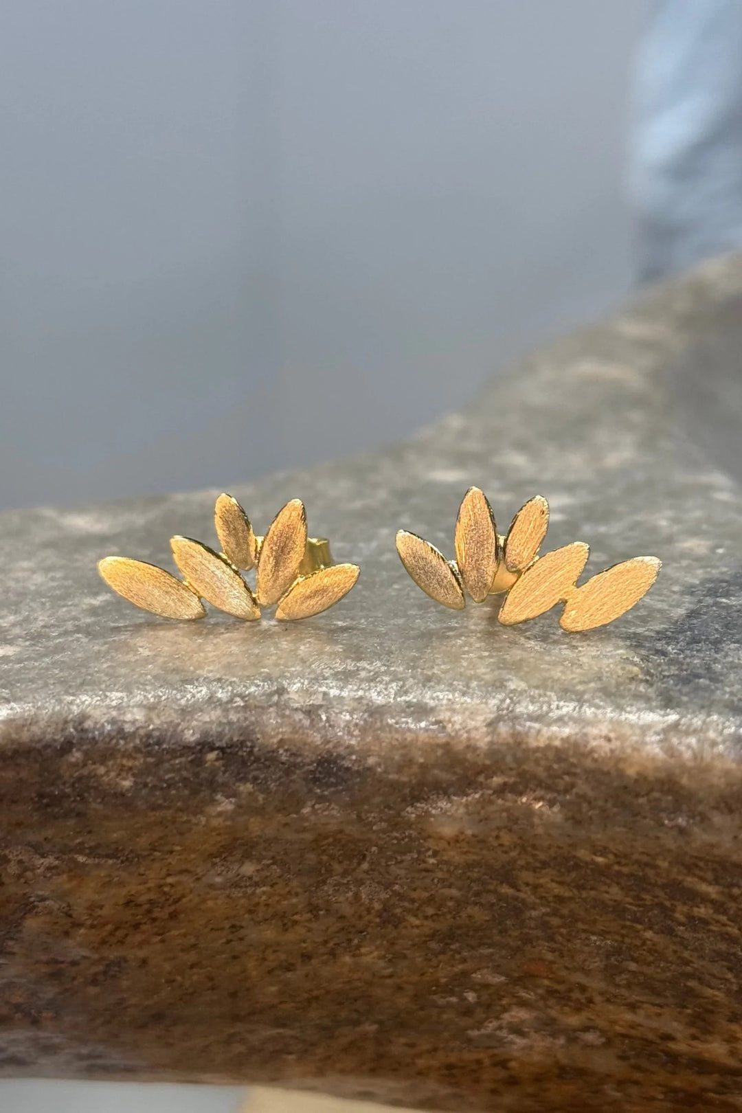 Guldbelagte øreringe med blomsterdesign, overlappende blade og tekstur på en stenet overflade.