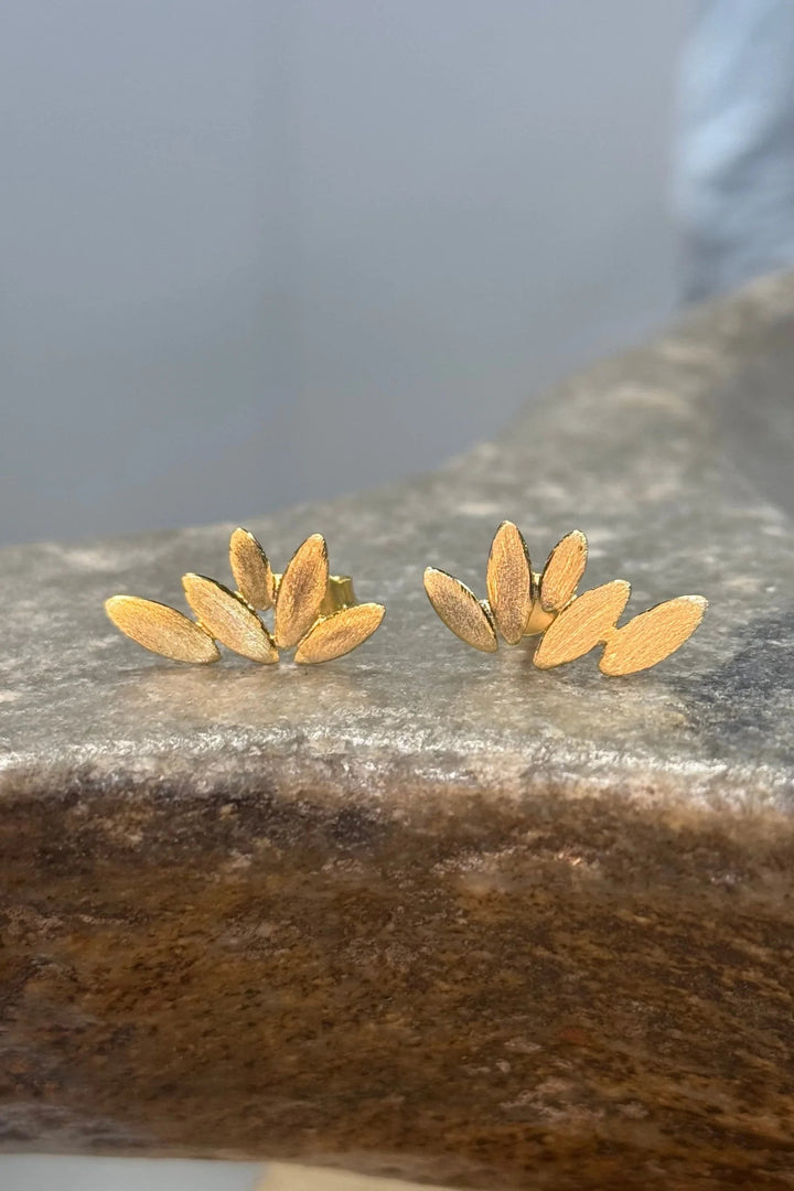 Guldbelagte øreringe med blomsterdesign, overlappende blade og tekstur på en stenet overflade.