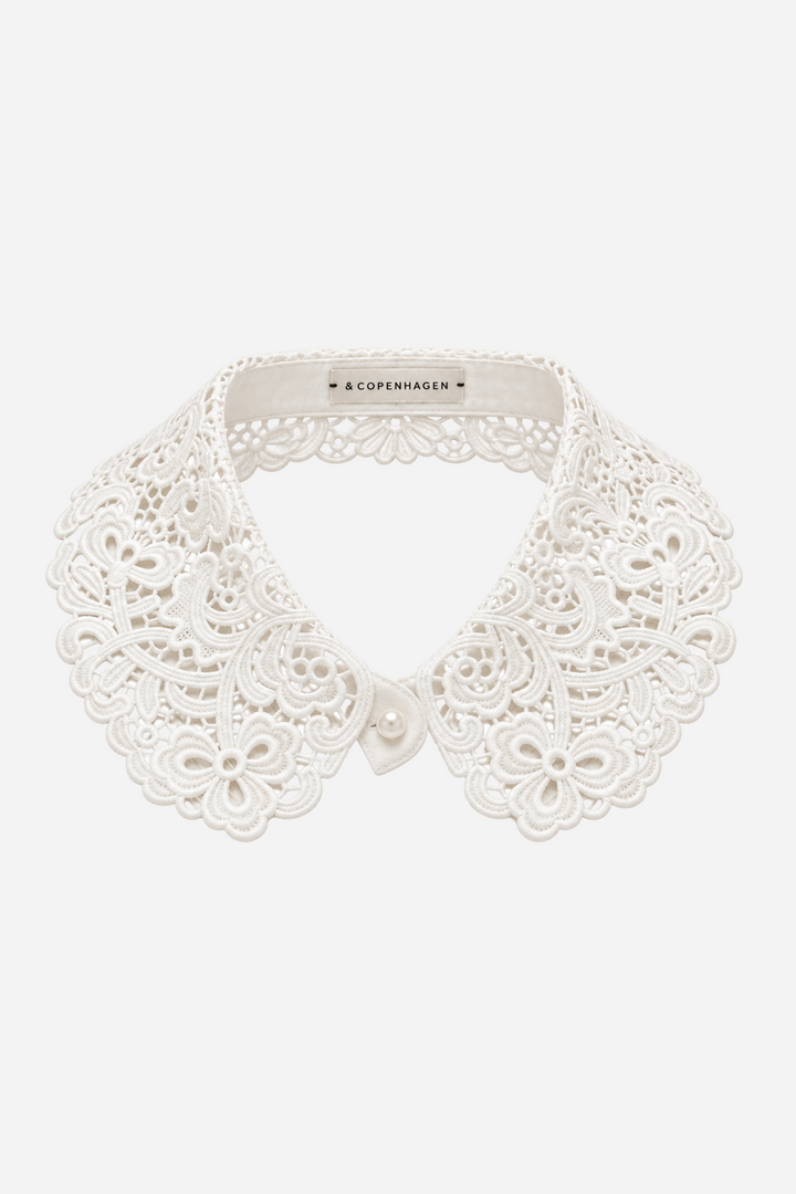 Hvid Darby blondekrave fra Andcopenhagen med detaljeret blomstermønster, dekorative udskæringer og bølgede kanter. Aftagelig krave med skjult knaplukning foran.
