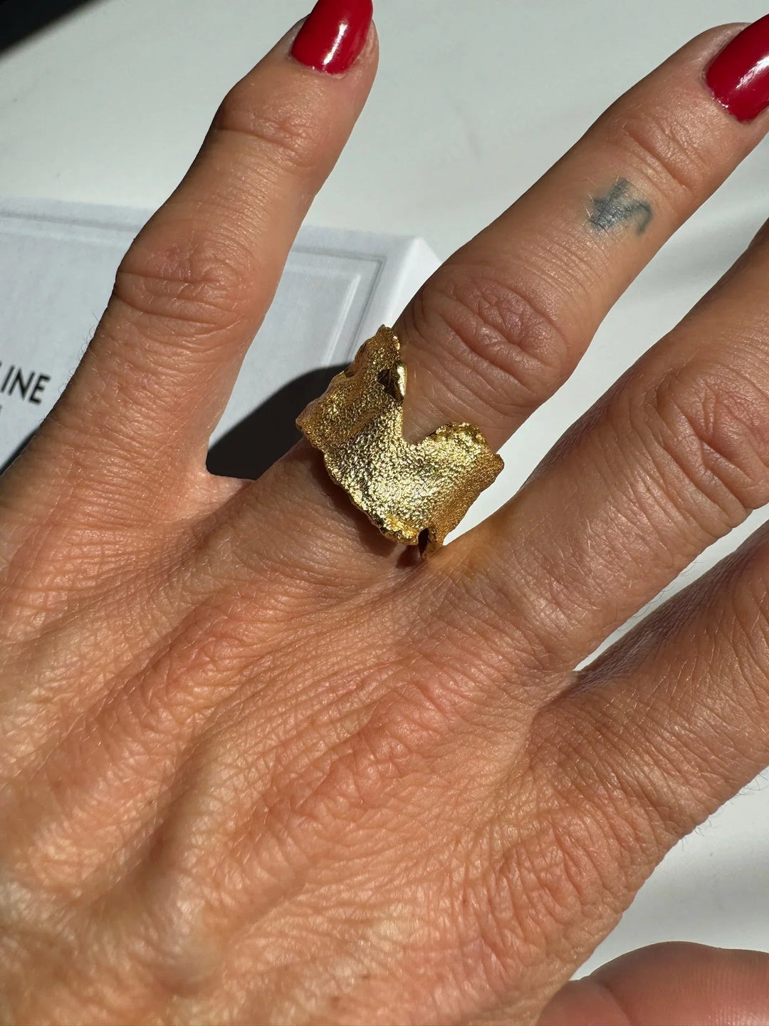 Hånd med rød neglelak bærer en guldbelagt ring med ujævn, organisk form.