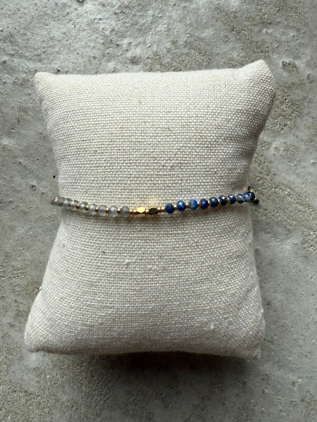 Perlearmbånd med blå, grå og gyldne perler på en tekstureret pude.