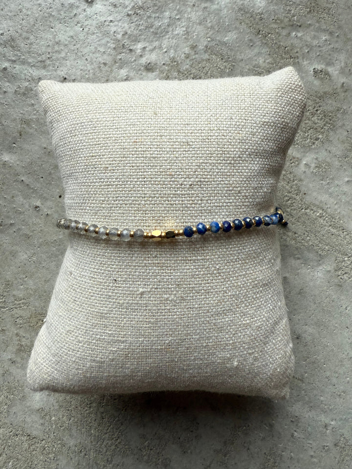 Perlearmbånd med blå, grå og gyldne perler på en tekstureret pude.