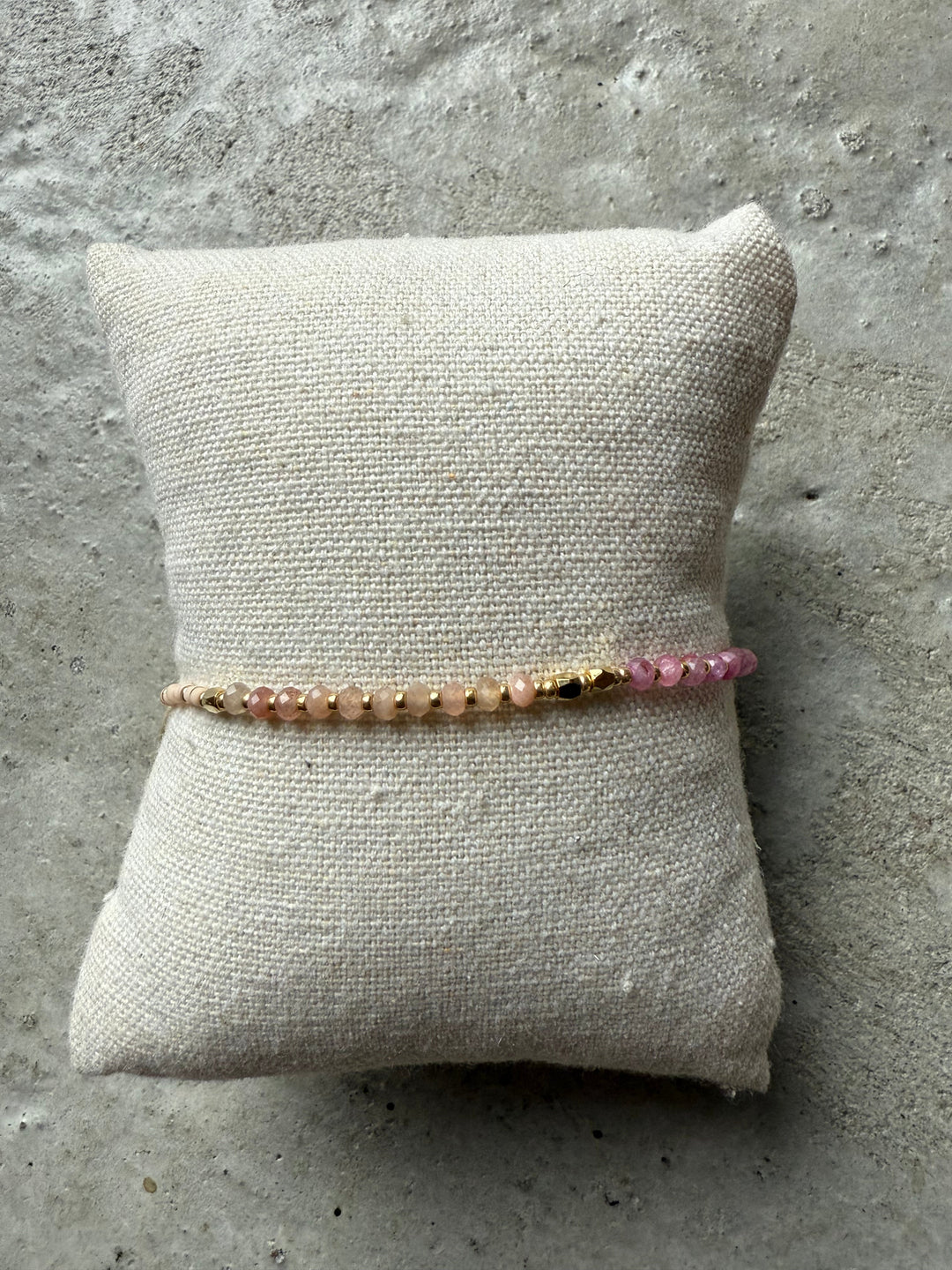 Perlearmbånd i rosa nuancer med gyldne detaljer, præsenteret på en beige pude mod grå baggrund.