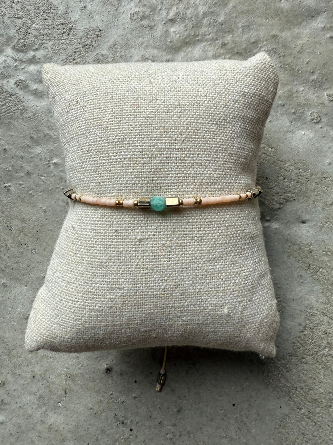 Armbånd med lyserøde perler, guldaccenter og en blå perle, præsenteret på en lys pude.