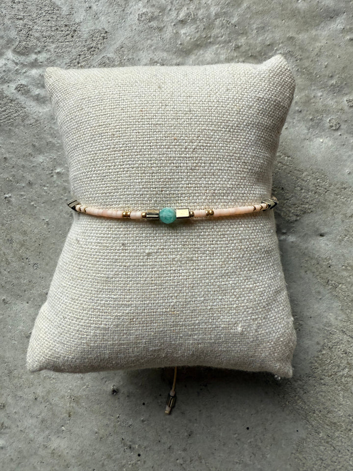 Armbånd med lyserøde perler, guldaccenter og en blå perle, præsenteret på en lys pude.