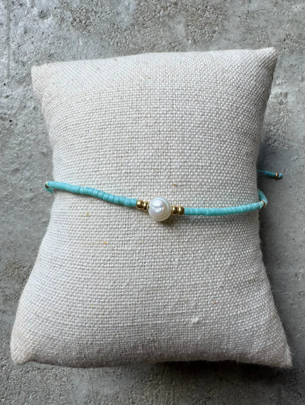 Turkis armbånd med central perle og guld accenter på lys pude.