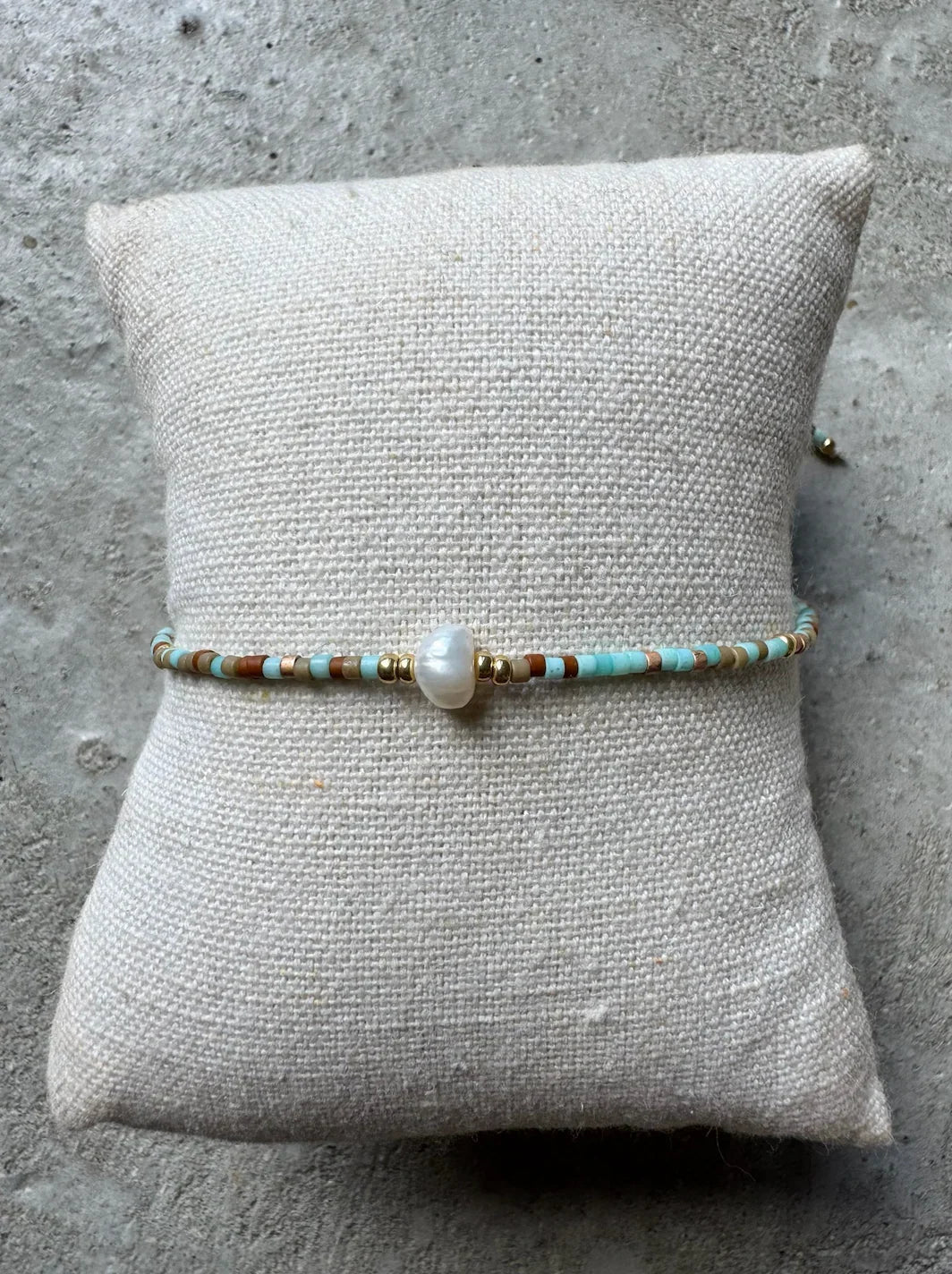 Armbånd med blå, brune og khaki perler og en central perle, lagt på en tekstureret pude.