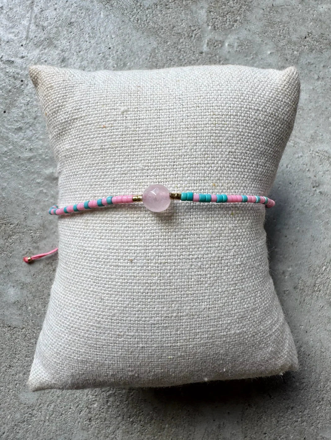 Perlearmbånd i pink og lyseblå med central lys perle, placeret på en beige pude.