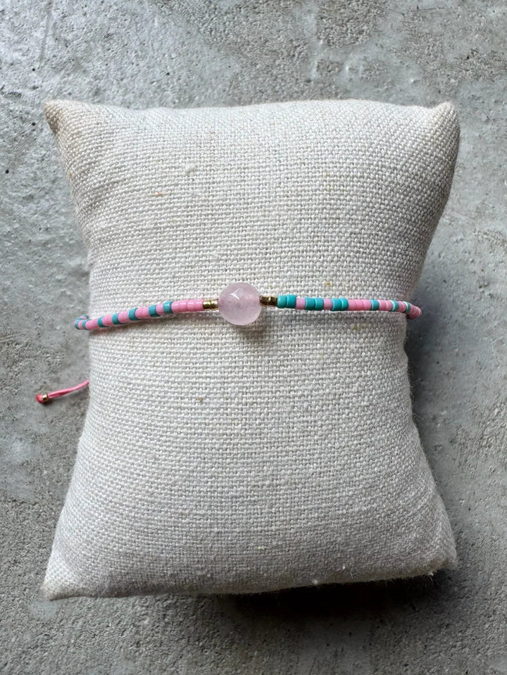 Perlearmbånd i pink og lyseblå med central lys perle, placeret på en beige pude.