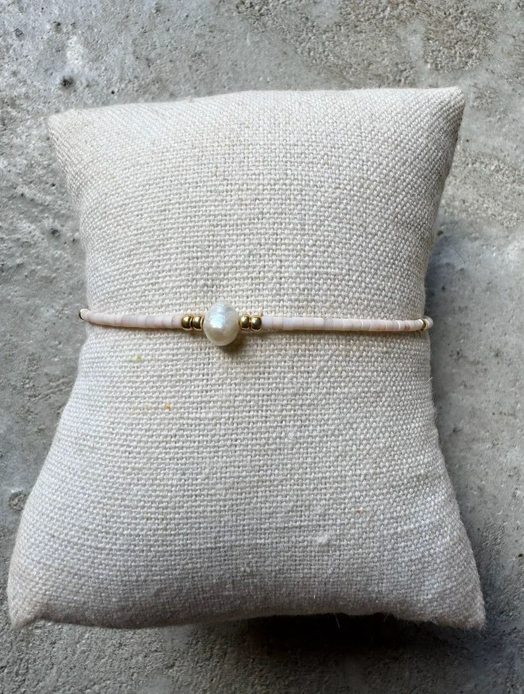 Beige perlearmbånd med central perle og gulddetaljer på tekstureret pude.