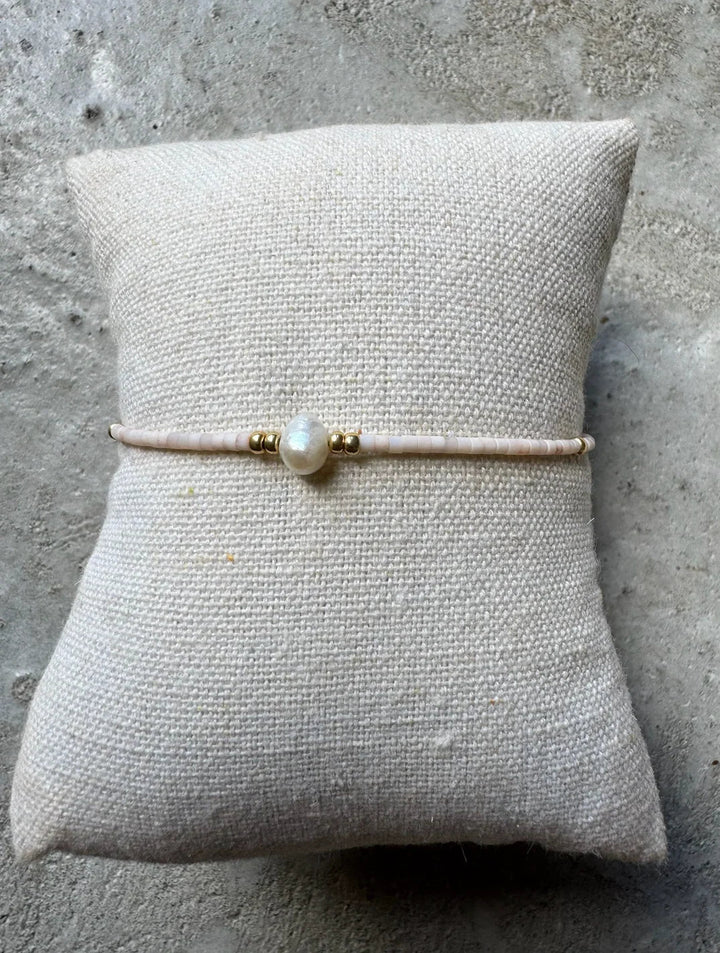 Beige perlearmbånd med central perle og gulddetaljer på tekstureret pude.