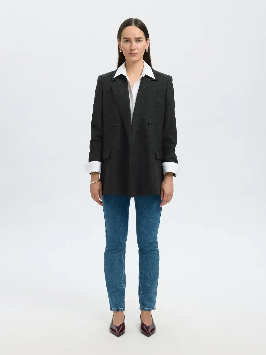 Kvinde i sort dobbeltknappet blazer med mønster, hvid skjorte, blå jeans og bordeaux sko.